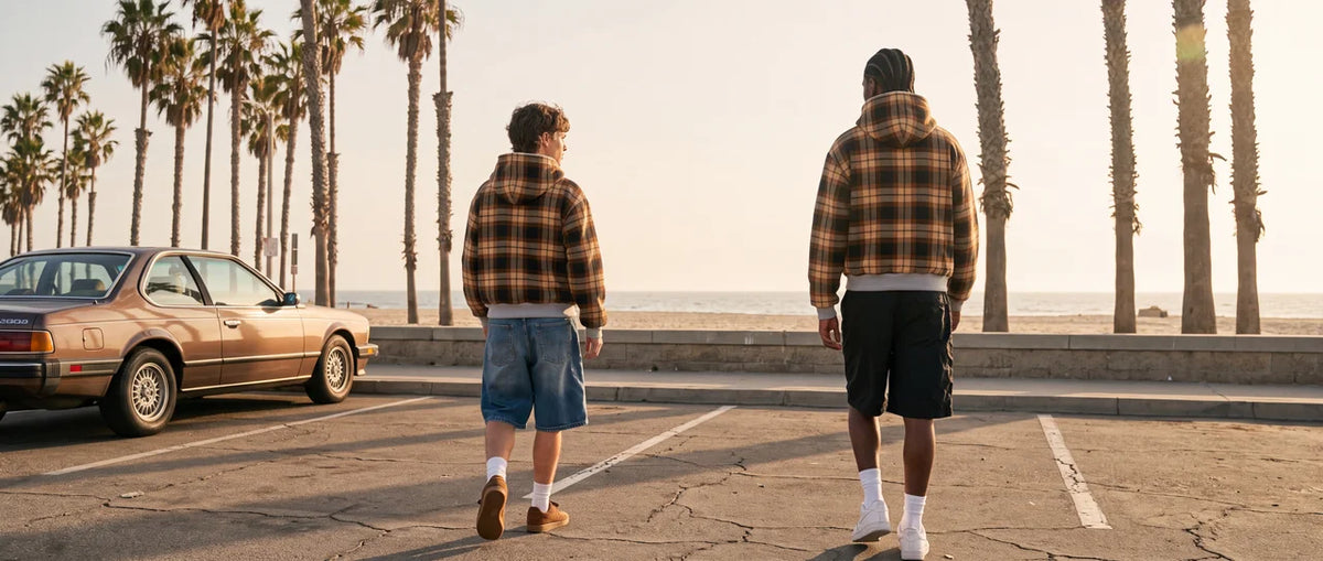 Two people walking along a palm-lined beachfront in ELEGANT plaid hooded jackets
